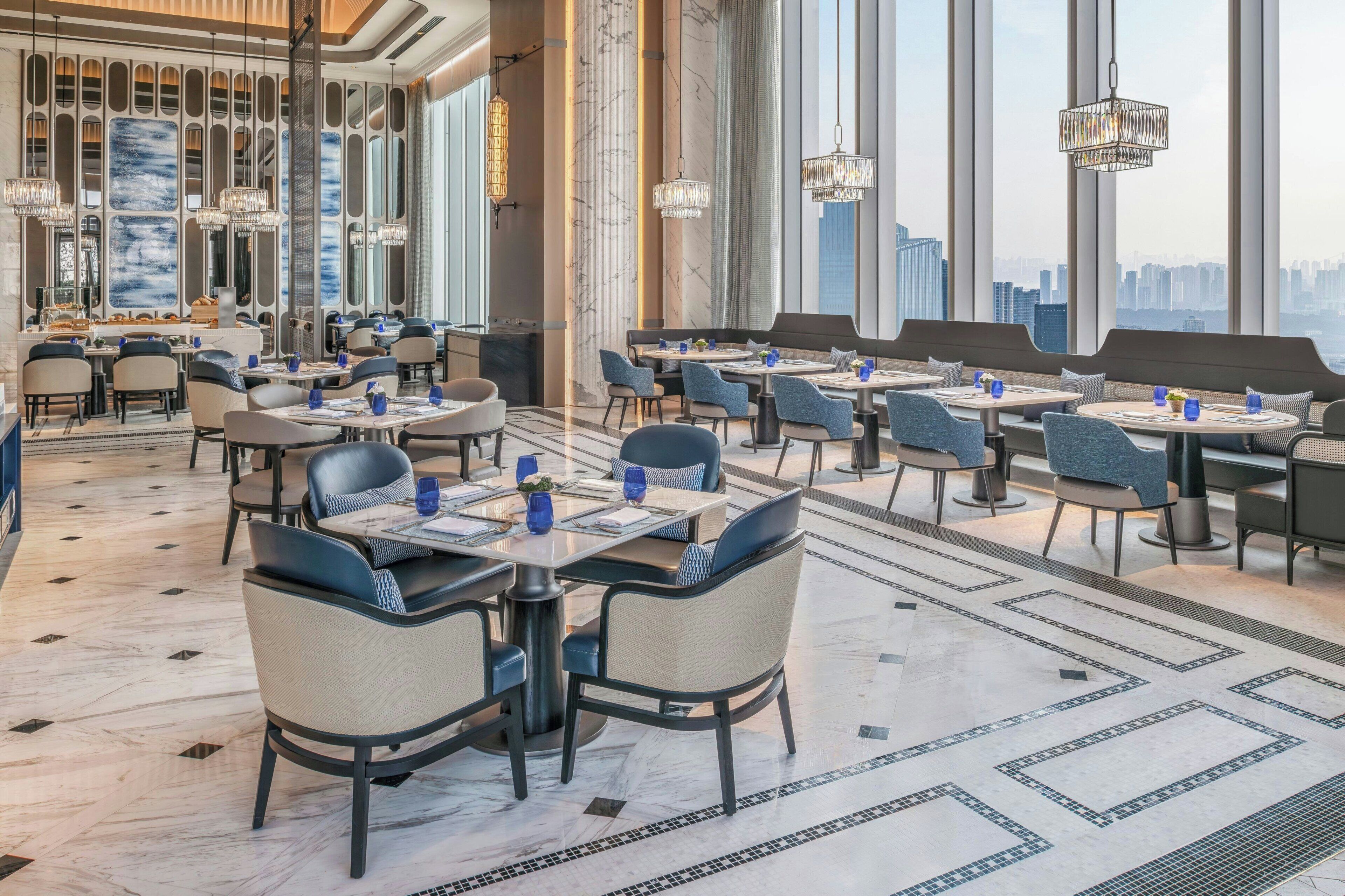 Un comedor espacioso con varias mesas y sillas, ventanas amplias con vista a la ciudad y lámparas modernas.