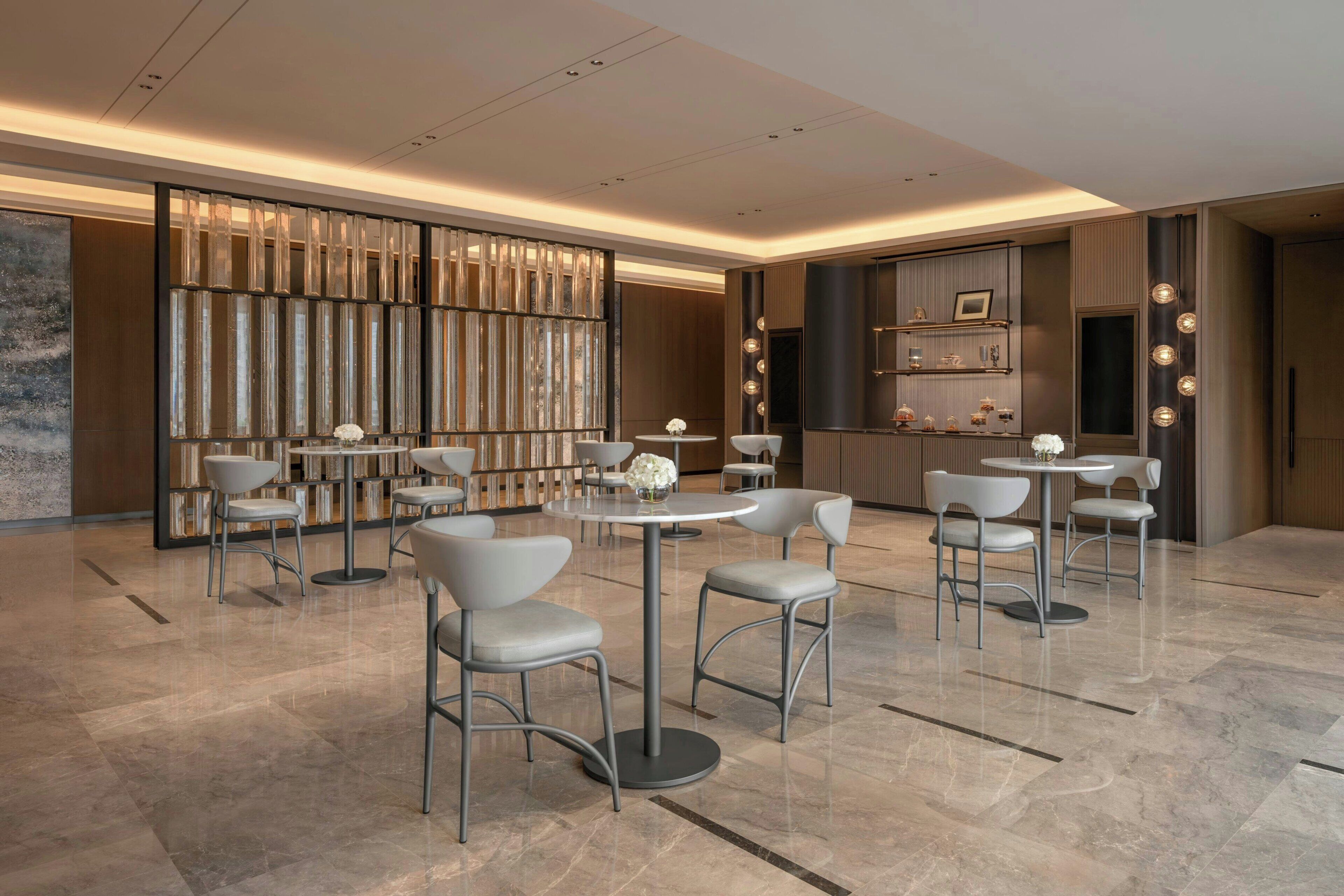Un interior moderno con varias mesas y sillas redondas, una pared decorativa con elementos verticales y una zona de cocina con estantes.