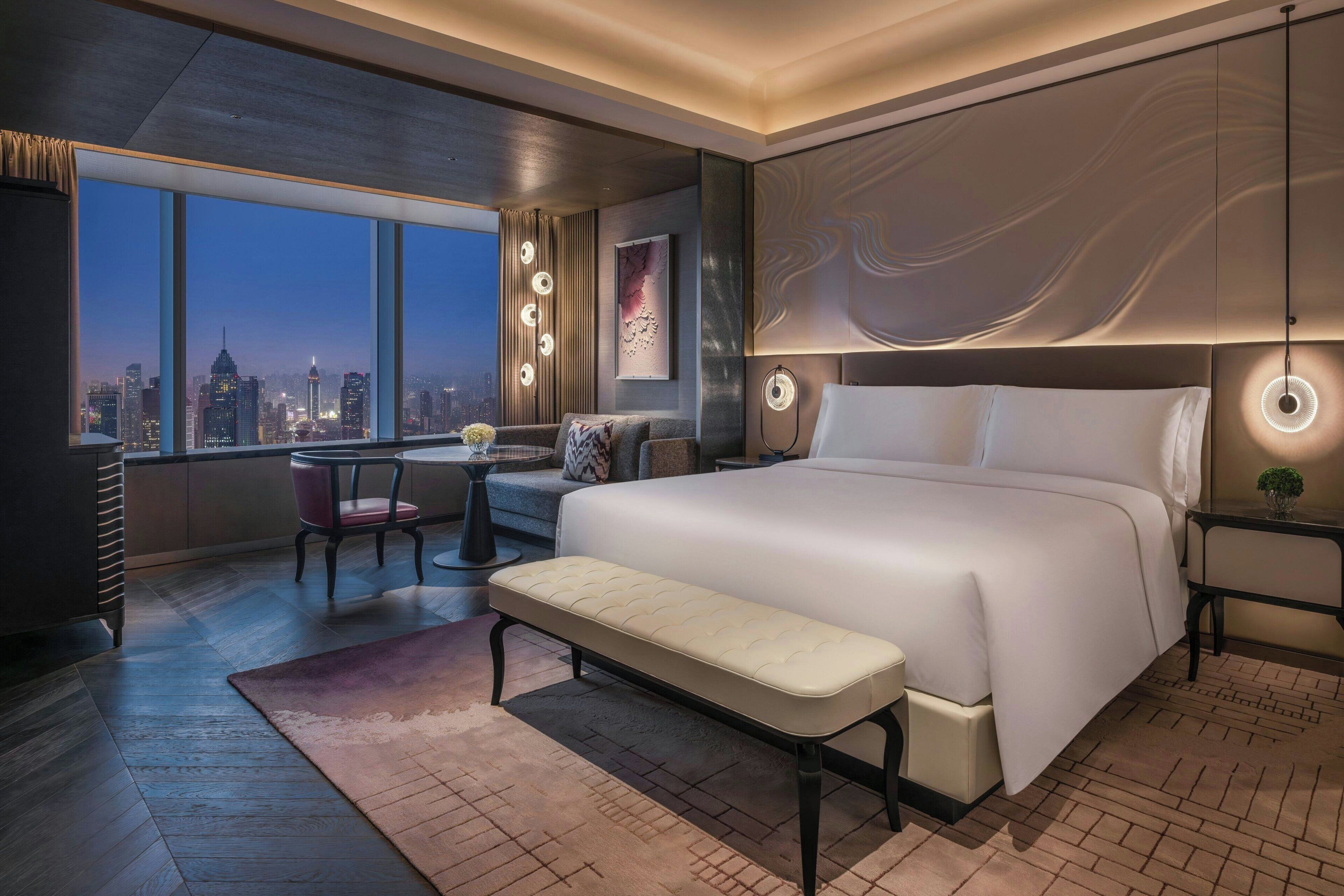 Una habitación de hotel moderna con una cama grande, un banco, una mesita pequeña y vistas del horizonte de la ciudad.