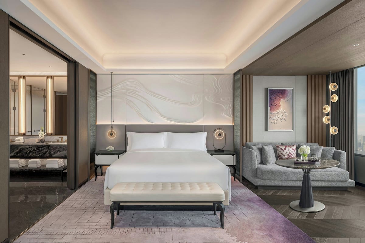 A modern hotel room with a large bed, a sofa, a small table, and a bathroom visible through an open door.