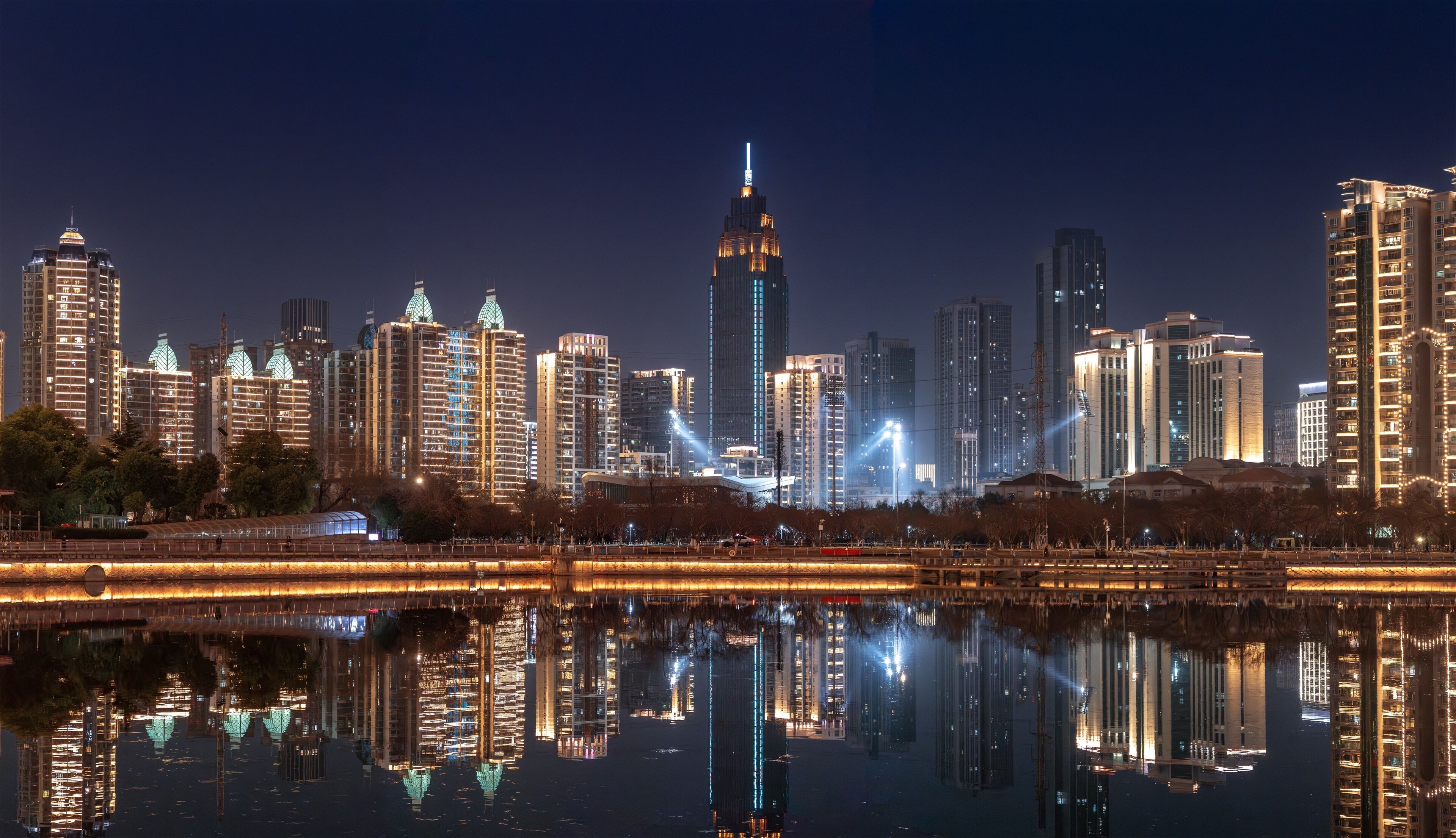 A nighttime cityscape with high-rise buildings reflected in a body of water.