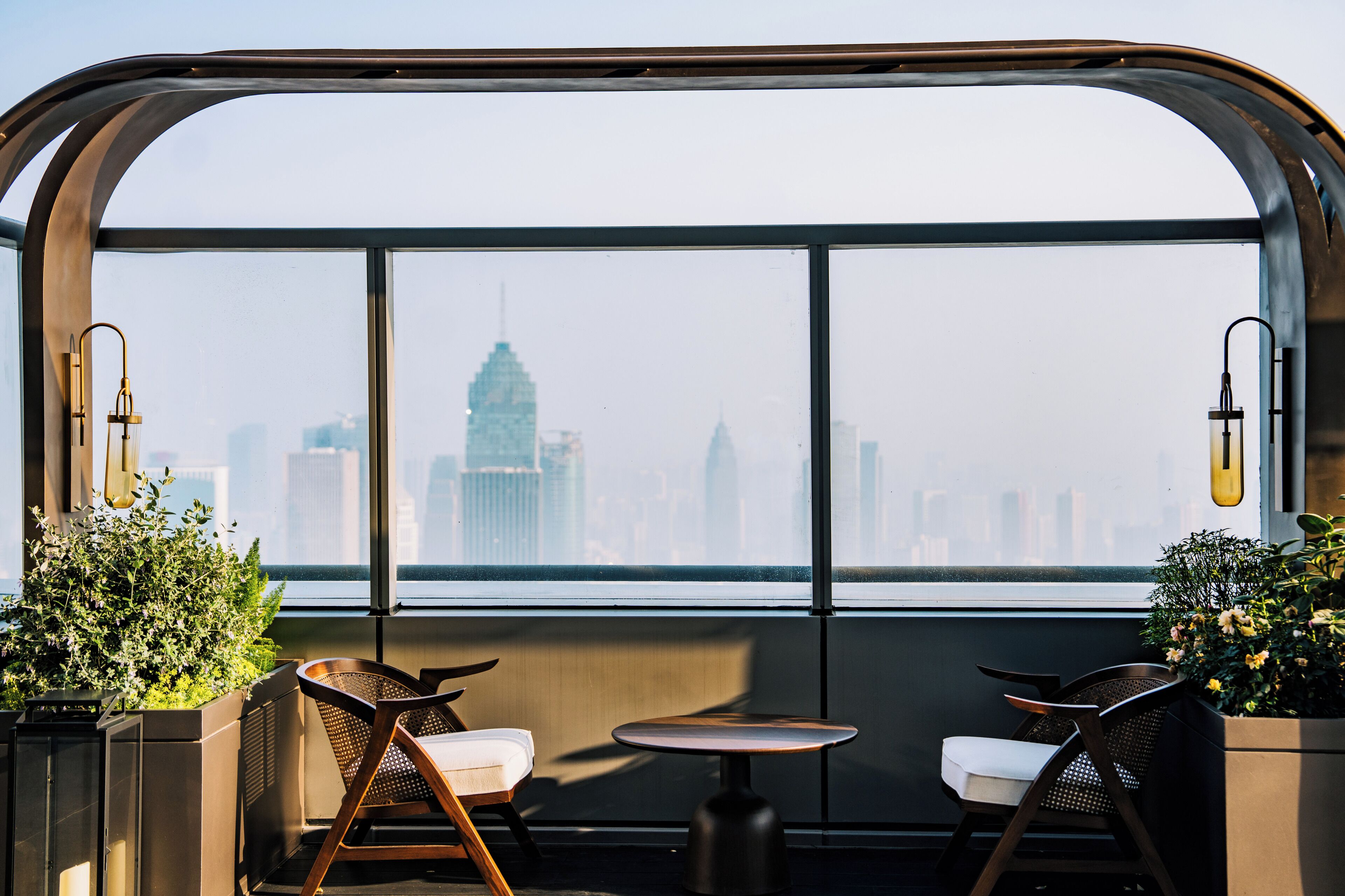 A high-rise interior with a panoramic view of a city skyline, featuring a seating area with chairs and a small table.
