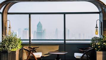 A high-rise interior with a panoramic view of a city skyline, featuring a seating area with chairs and a small table.