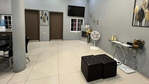 A spacious living area with a tiled floor, a black ottoman, a standing fan, a flat-screen TV mounted on the wall, and a small table with decorative items.