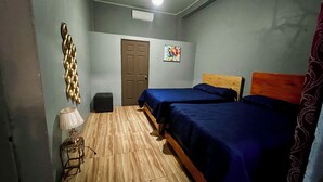 Ein kleiner, gut beleuchteter Raum mit zwei Betten, Holzboden, einer Tür, einem kleinen Tisch mit Lampe und einer Wanddekoration.