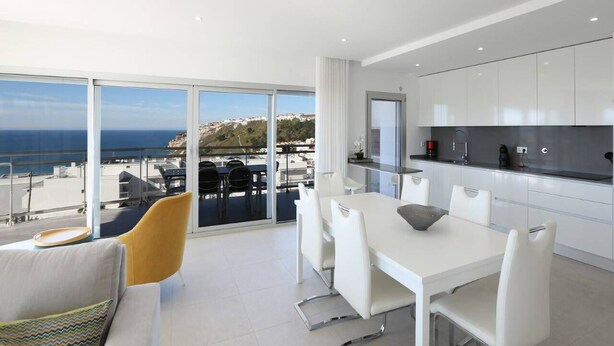 A modern kitchen with white cabinets and a dining area with a white table and chairs. There is a large window with a view of the ocean and a balcony with outdoor seating. - Aziadé Horizons - Ocean View (Nazaré)