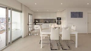 A modern kitchen with white cabinets, a dining area with a white table and chairs, and a large sliding glass door leading to a balcony. - Aziadé Horizons - Ocean View (Nazaré)