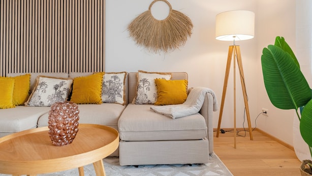 Un salon moderne avec un canapé d’angle, une table basse ronde en bois, un lampadaire et une grande plante verte.