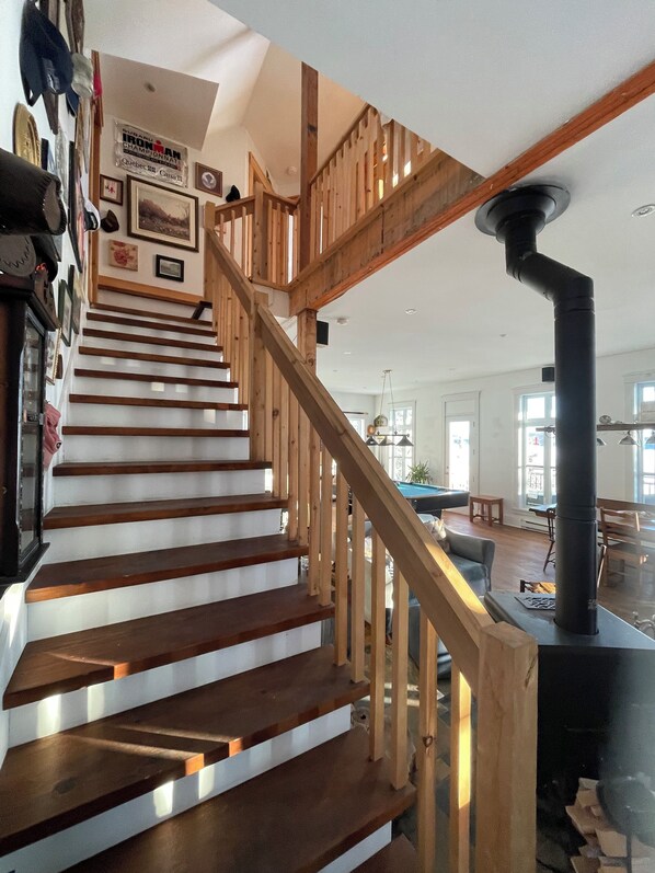 Una escalera de madera con una tubería negra que recorre la pared, que conduce a una sala de estar con mesa de billar y chimenea.