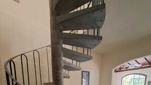 Un escalier en colimaçon avec une rampe en métal, un banc en bois et un canapé dans une pièce dont le sol est en pierre et qui possède une fenêtre.