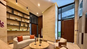 Una sala de estar moderna con un sofá curvo, una mesa de centro redonda y sillas de madera. Se observan estanterías con objetos decorativos y un amplio ventanal que proporciona luz natural.