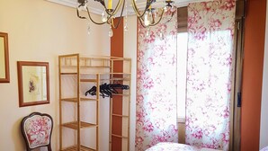 Ein Schlafzimmer mit zwei Betten, einem Holzschrank, einem Sessel, einer Deckenleuchte und blumigen Vorhängen.