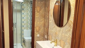 Ein Badezimmer mit weißem Waschtisch, goldenem Wasserhahn und einem Spiegel mit goldener Rahmung. Es gibt eine Duschkabine mit Glastür und einer gefliesten Wand.