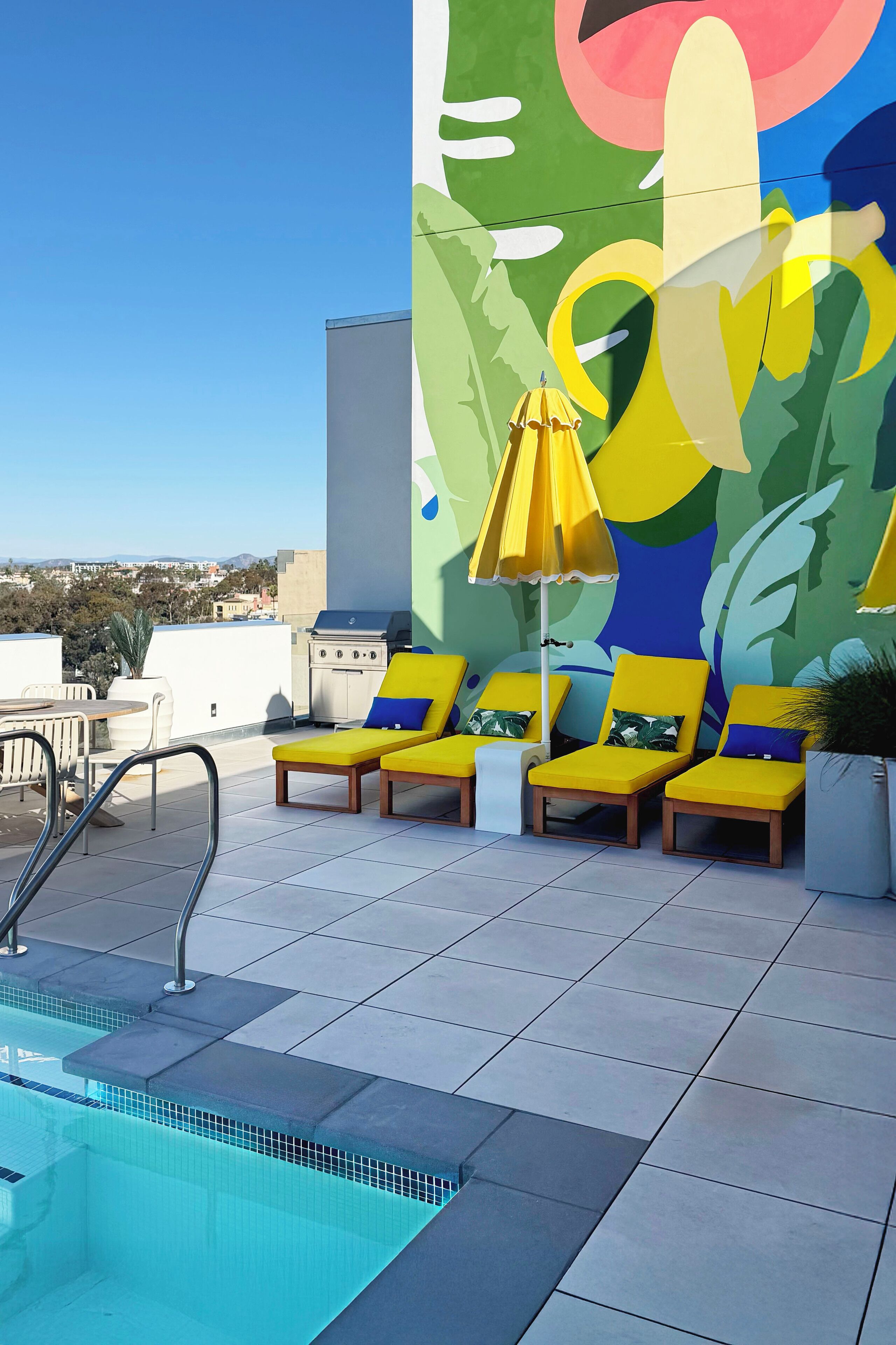 A rooftop pool area with a colorful mural, yellow lounge chairs, and a grill.