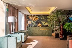 A reception area with a green reception desk, a seating area with light green sofas, a large potted plant, and a wall with a tropical leaf mural. - AMANA Hotel Airport  (Tan Binh)