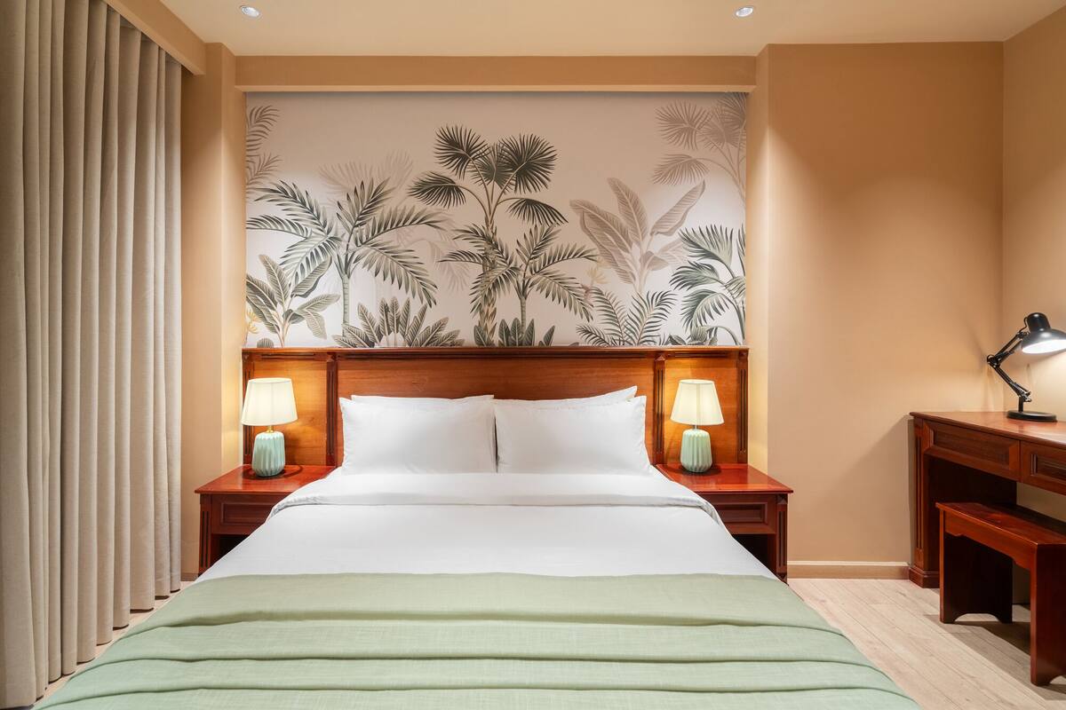 Une chambre d’hôtel avec un grand lit, deux tables de chevet avec des lampes, une tête de lit en bois et un papier peint à motifs tropicaux.