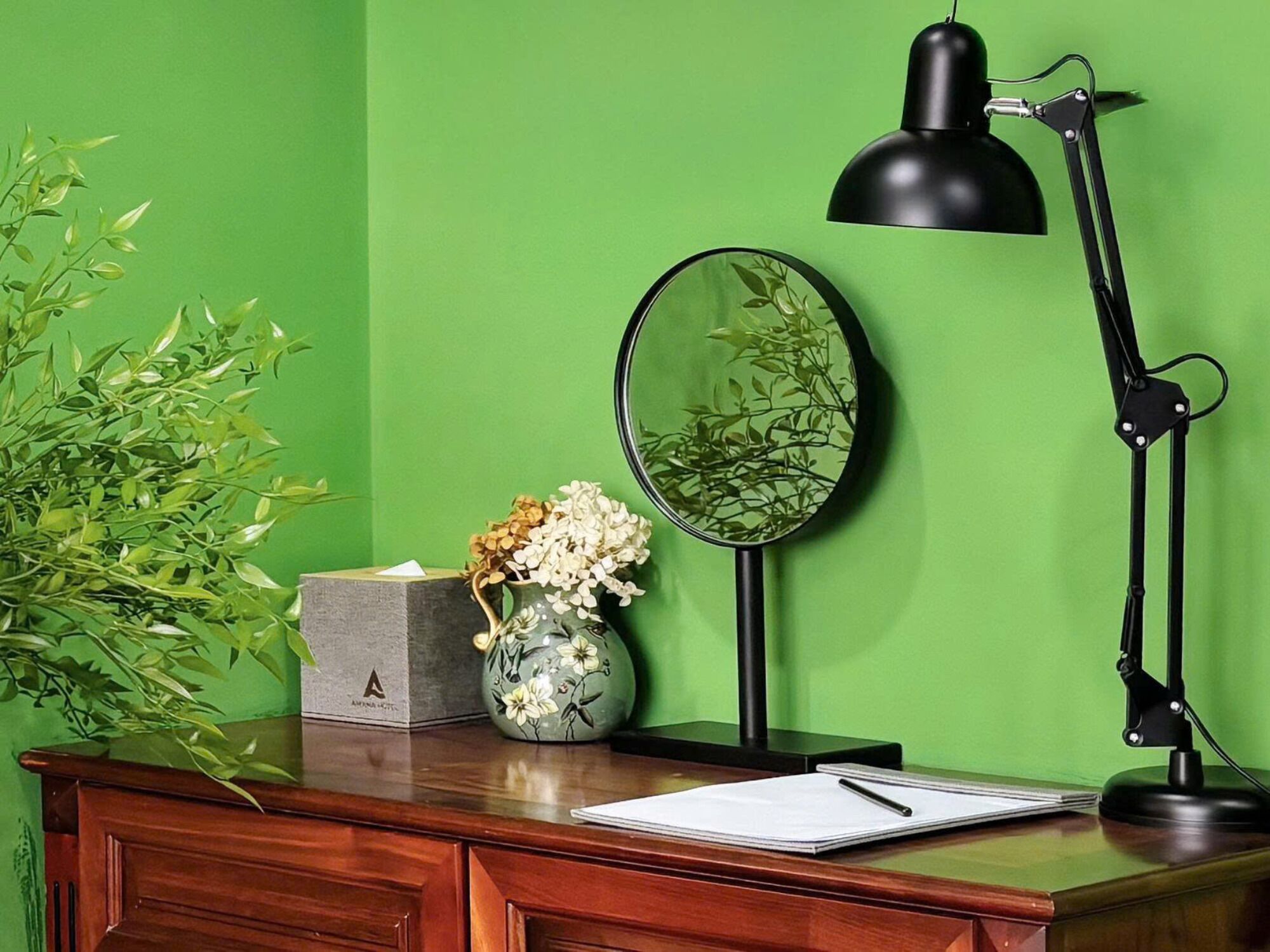 Un escritorio de madera con un jarrón de flores, un espejo y una lámpara de escritorio negra frente a una pared verde.