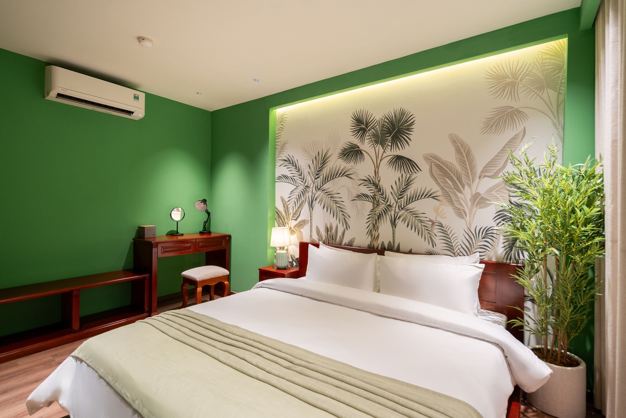 A hotel room with a bed, a desk, a chair, a lamp, and a potted plant.