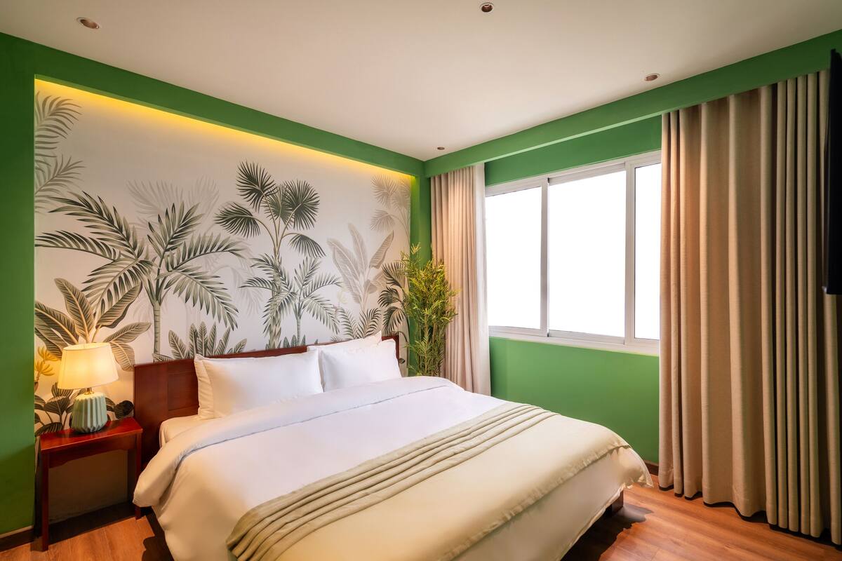 Un lit bien fait, avec du linge de lit blanc, un mur d’accent vert orné d’un motif de feuilles tropicales, une table de chevet en bois avec une lampe, et une grande fenêtre avec des rideaux beige.