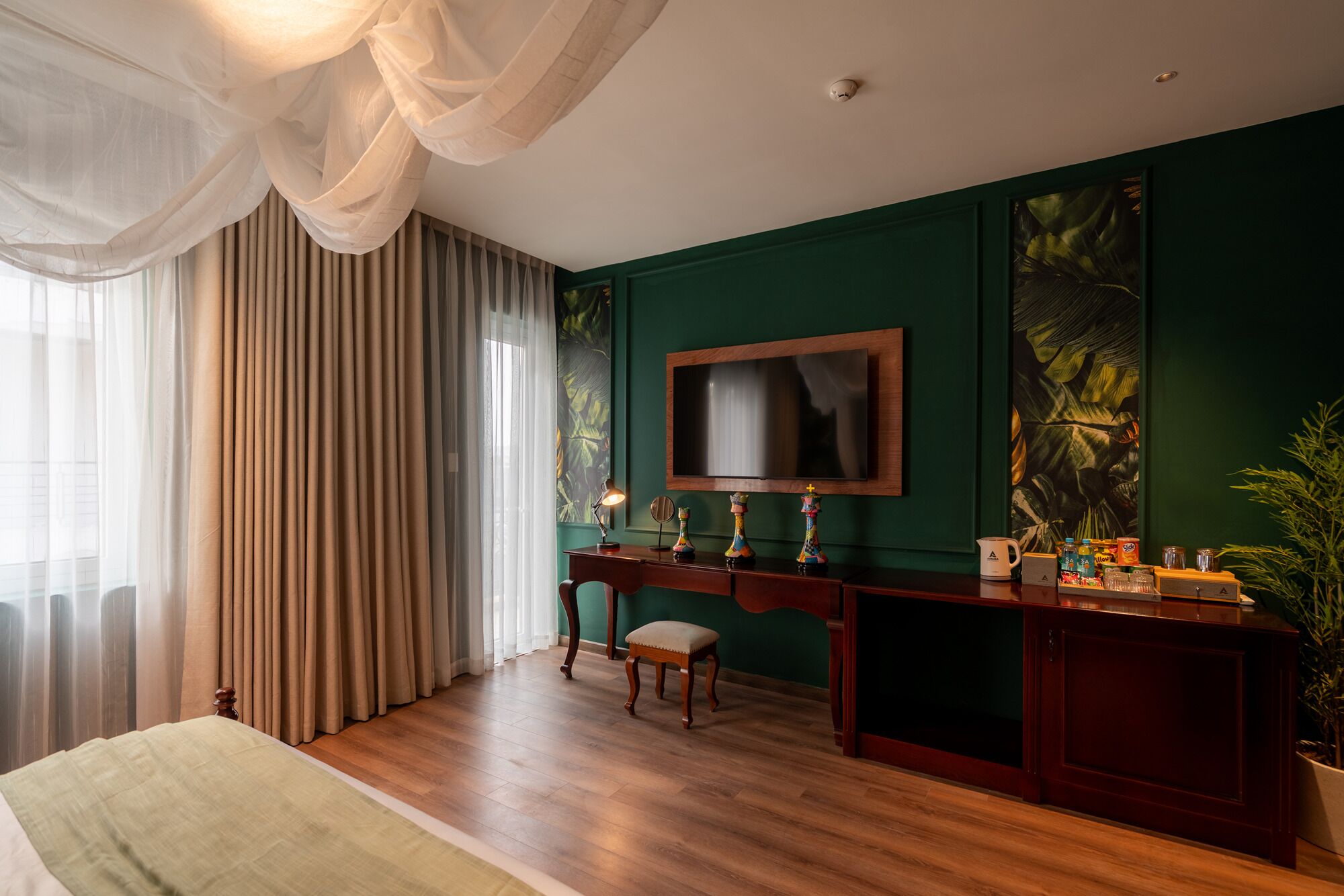 A hotel room with a large bed, a desk with a flat-screen TV, a potted plant, and a window with curtains.