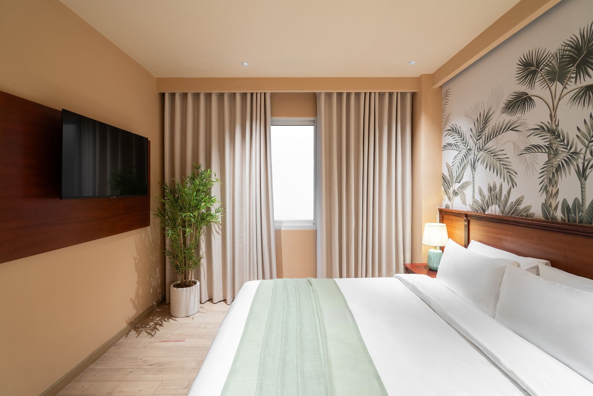 Una habitación de hotel con una cama grande, una planta en maceta, un televisor montado en la pared y un papel tapiz tropical.