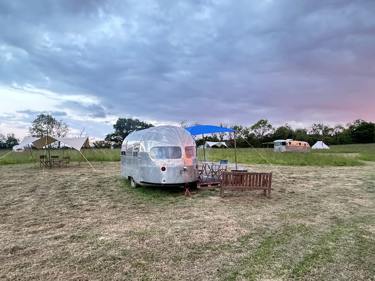 A vintage silver trailer with a blue canopy, set in a grassy field with other camping setups in the background.