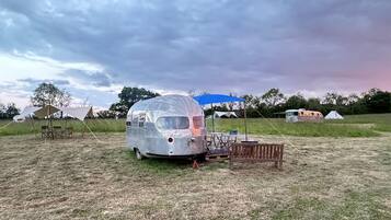 A vintage silver trailer with a blue canopy, set in a grassy field with other camping setups in the background.