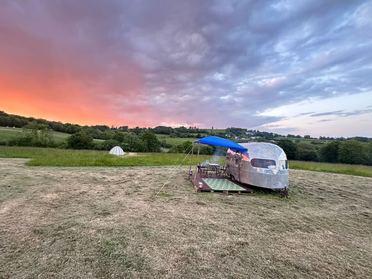 A silver RV with a blue canopy, a picnic table, and a grill set up in a grassy field with a sunset in the background.