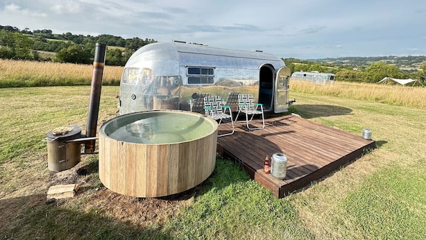 A vintage trailer with a hot tub and wooden deck, surrounded by a grassy field.