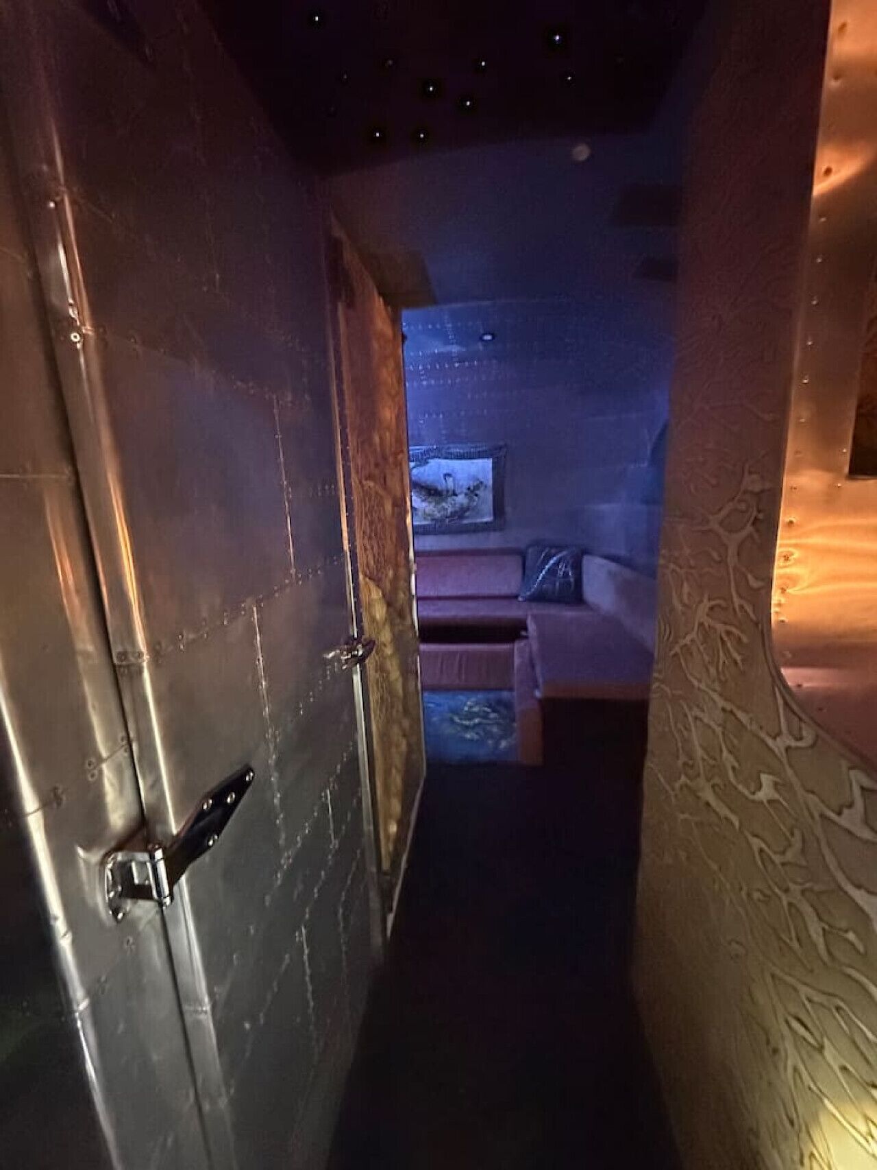 Un couloir étroit mène à une pièce où se trouvent un lit, une petite table et un téléviseur fixé au mur.