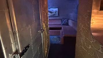 Un couloir étroit mène à une pièce où se trouvent un lit, une petite table et un téléviseur fixé au mur.
