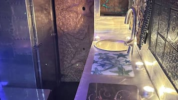Ein Badezimmer mit Marmorwand, Waschbecken und Spiegel. Ein kleines Regal zeigt eine Kerze und ein dekoratives Objekt.