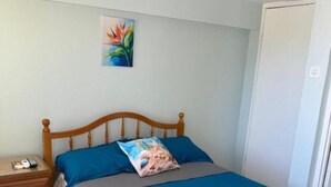 Ein Schlafzimmer mit einem hölzernen Bett, einem Nachttisch, einer blauen Bettdecke, einem Kissen mit floralem Muster und einem Gemälde an der Wand.