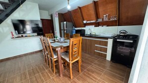 Una cocina con armarios de madera, una mesa de comedor con sillas, un refrigerador, una estufa y un televisor montado en la pared.