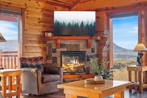 Ein Interieur einer Blockhütte mit Kamin aus Stein, einem darüber hängenden Fernseher und Bergblick durch große Fenster.