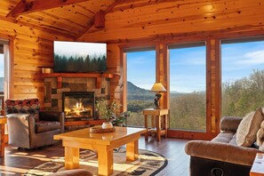 Ein Blockhaus-Interieur mit Kamin, einem darüber angebrachten Fernseher und einem großen Fenster mit Blick auf den umliegenden Wald.