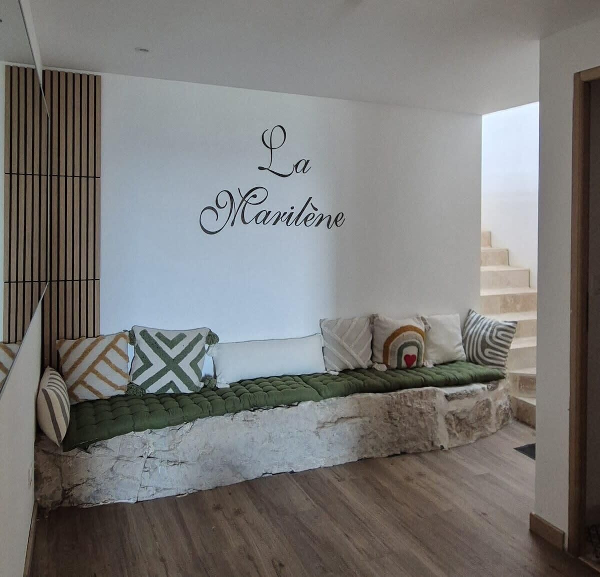 A cozy seating area with a stone bench and cushions, a wall with the text "La Maritene," and a staircase in the background.