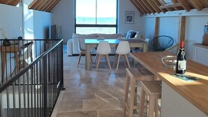 Een kamer onder het dak met een schuine kap, een eethoek met tafel en stoelen, een keuken met aanrecht en een groot raam met uitzicht.