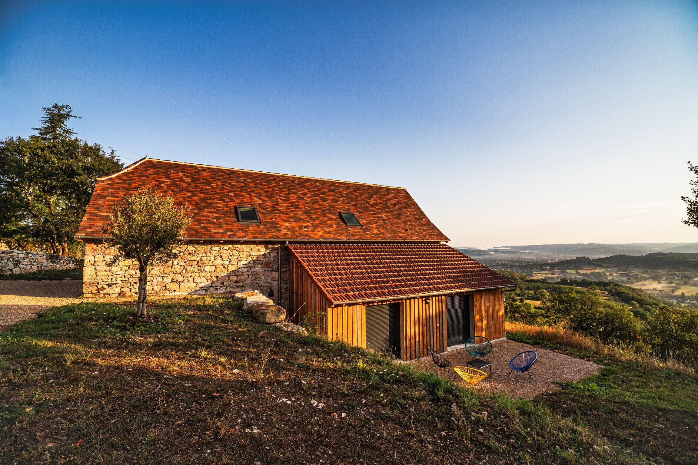 A rustic stone house with a sloped roof, a small wooden extension, and a view of the countryside.