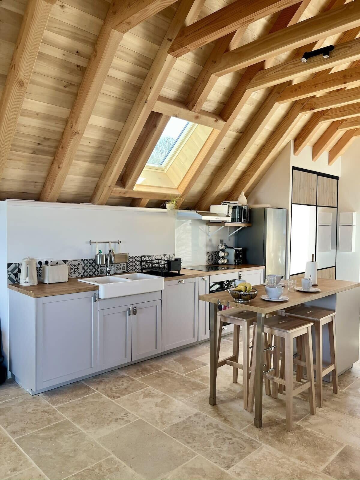 A modern kitchen with a sloped ceiling, wooden beams, a skylight, white cabinets, a wooden dining table, and a built-in oven.