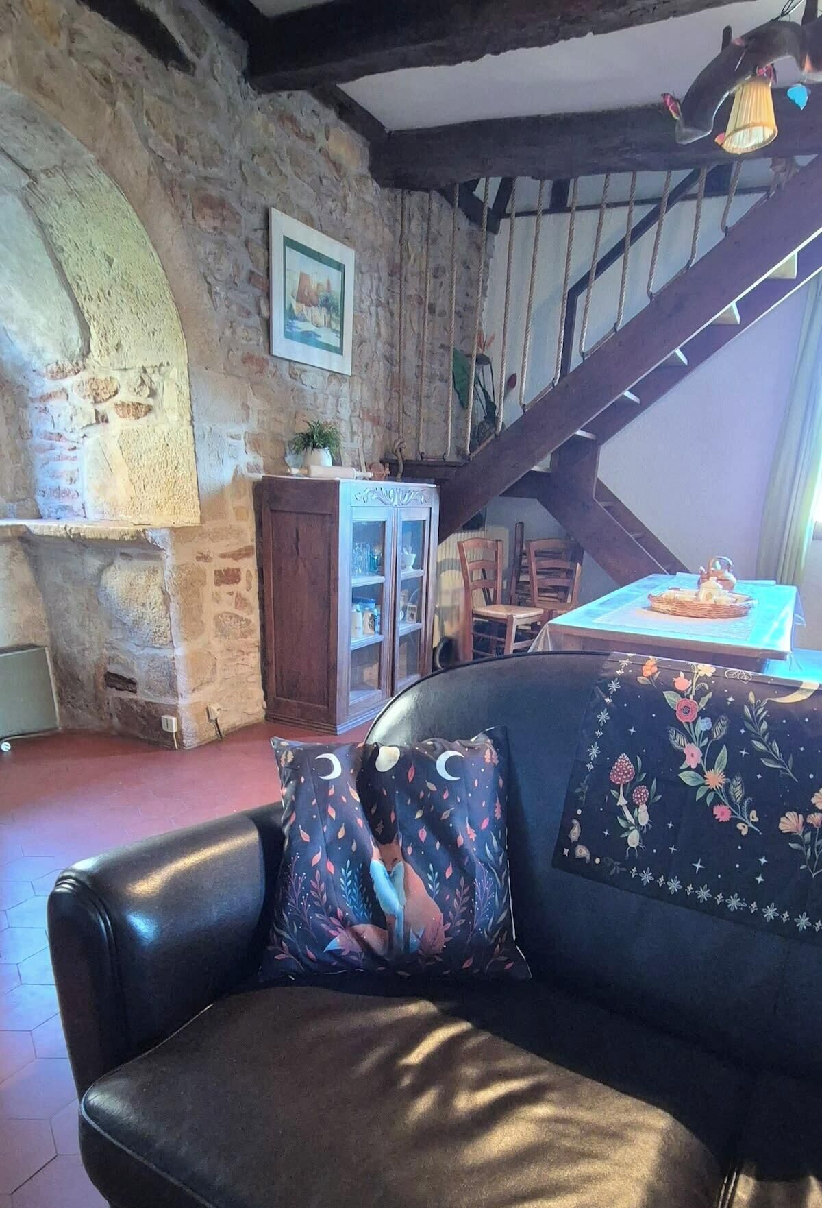 Un interior rústico con pared de piedra, una escalera de madera, un sofá de cuero con cojines estampados y una mesa con una cesta.