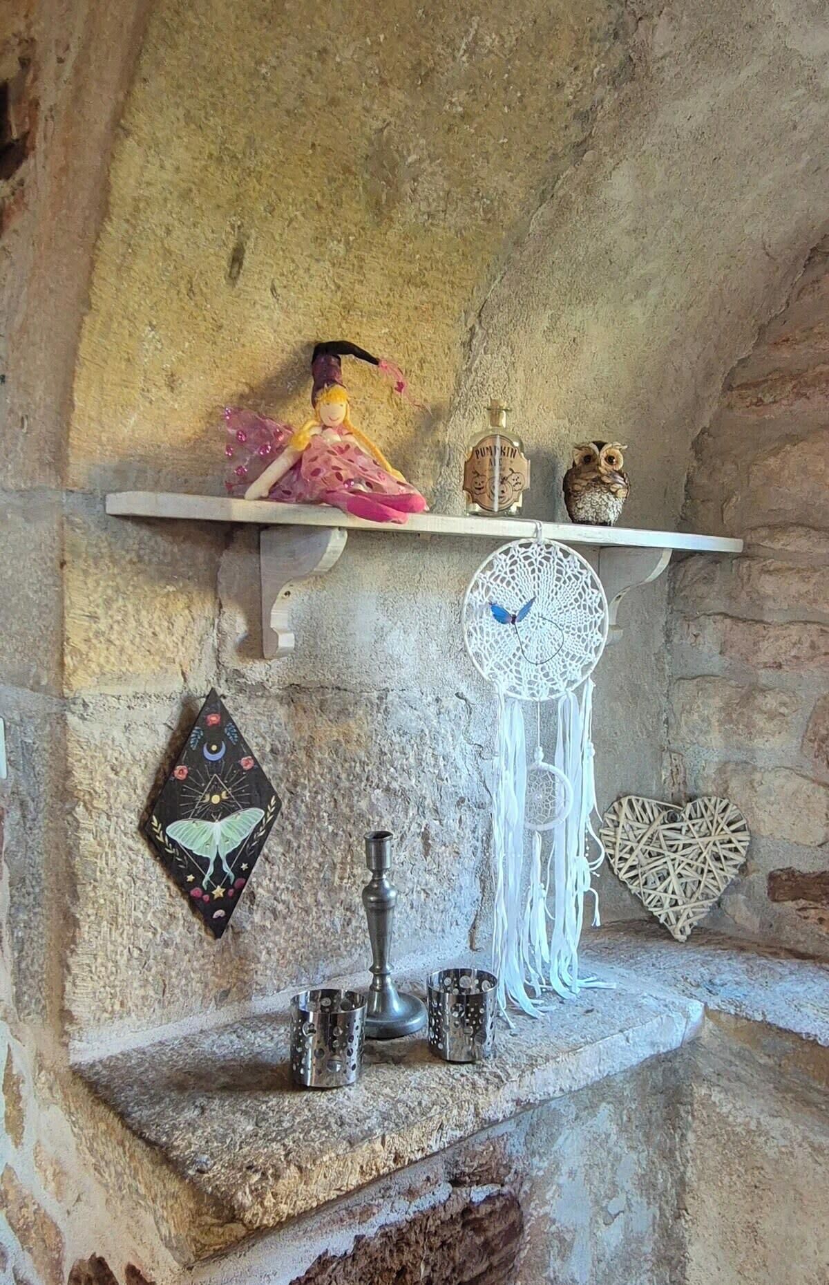 A stone wall with a shelf holding a doll, a bottle, and an owl. Below, there are dreamcatchers, a candle, and cups.