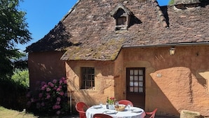 Une maison en pierre avec un toit en tuiles, une cheminée et une petite fenêtre. On y voit une table entourée de chaises, placée à l’extérieur, ainsi qu’un parterre de fleurs roses.