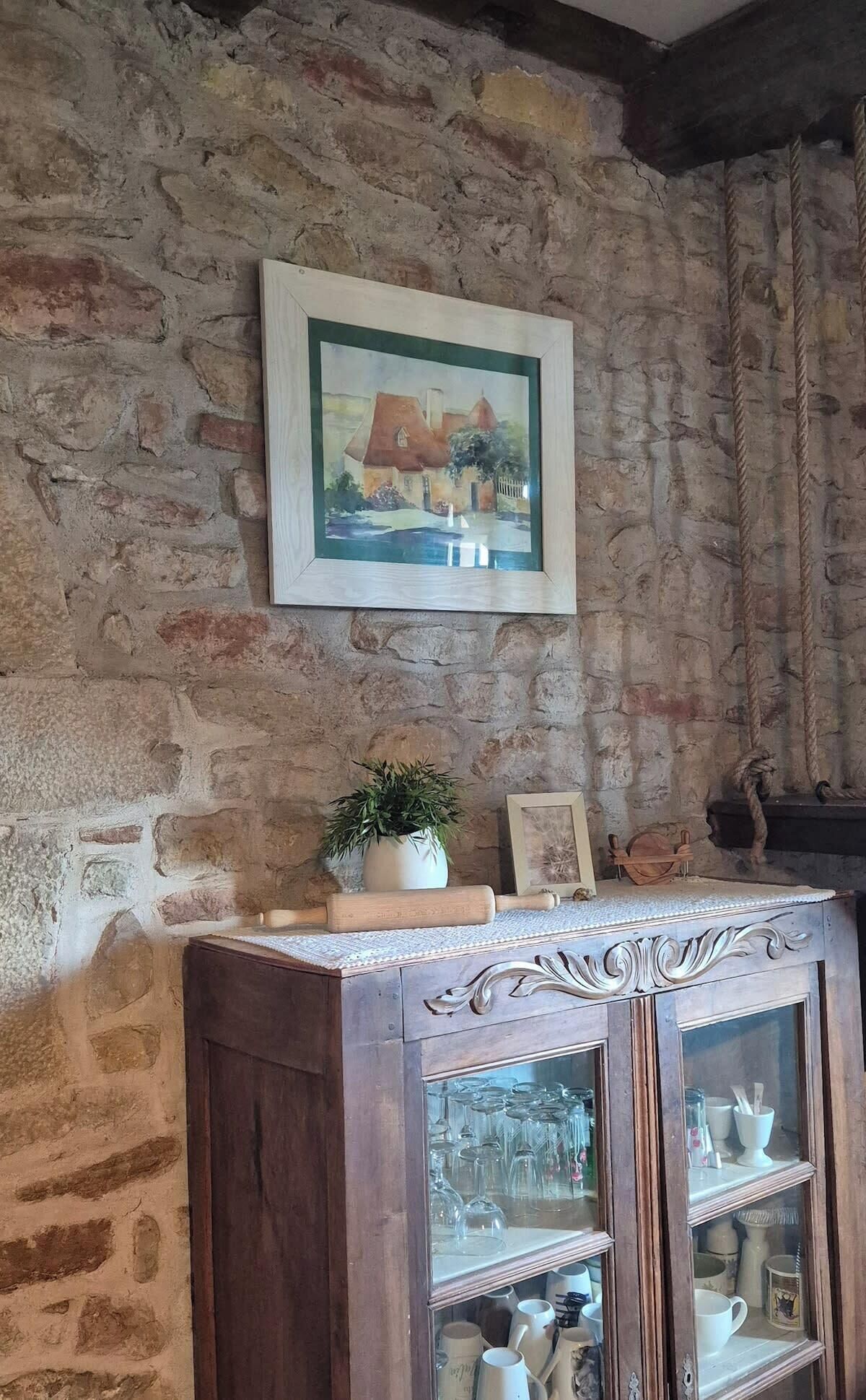 Ein rustikaler Raum mit Steinwand, einem gerahmten Gemälde eines Hauses, einer Holzhütte mit Glastüren und einem Blumentopf.
