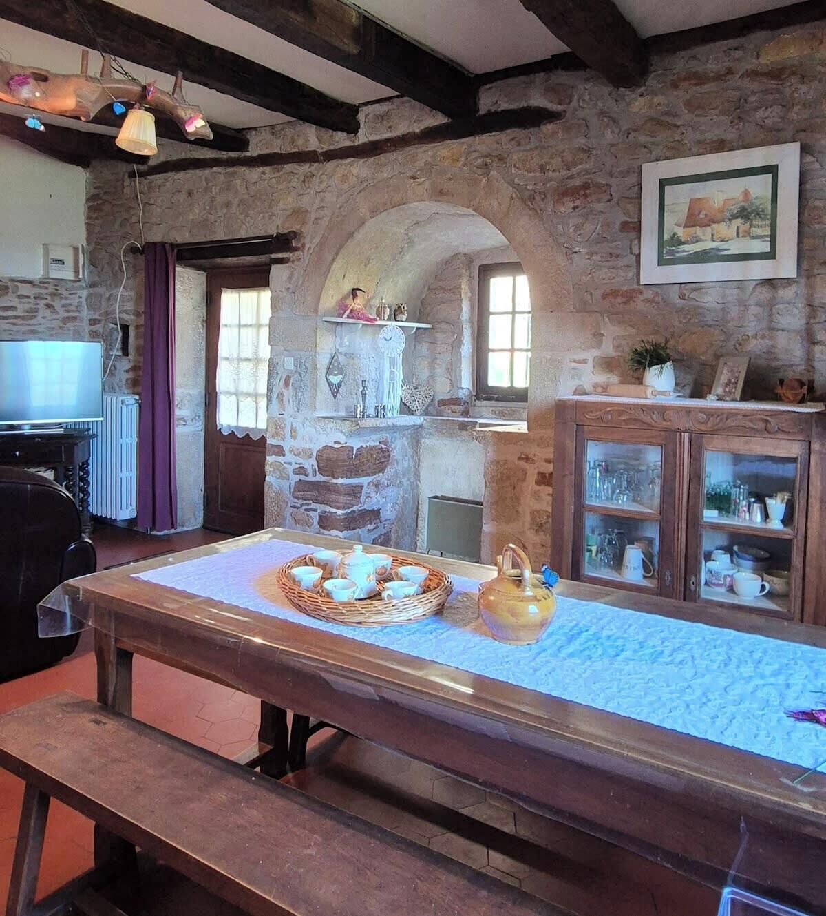 Una habitación rústica con pared de piedra, una mesa de madera con juego de té, un aparador con puertas de cristal y un cuadro en la pared.