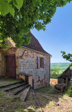 A rustic stone building with a steeply pitched roof, a wooden door, and a small porch area. - l'Ardoise, Maison au pied du château de Castelnau (Prudhomat)