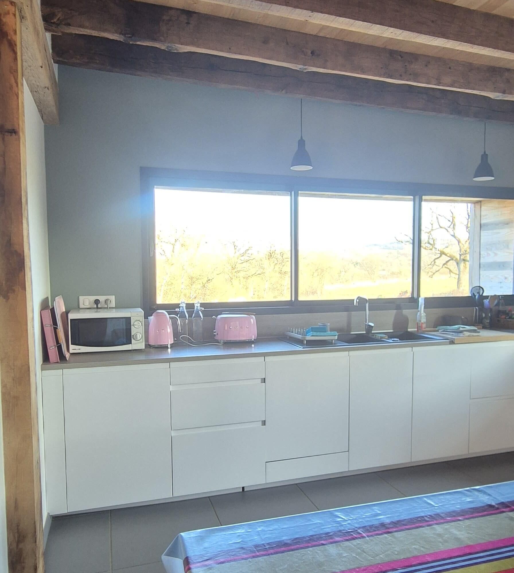 Una cocina con un ventanal grande, gabinetes blancos, un microondas, dos tostadoras rosadas y lámparas colgantes.