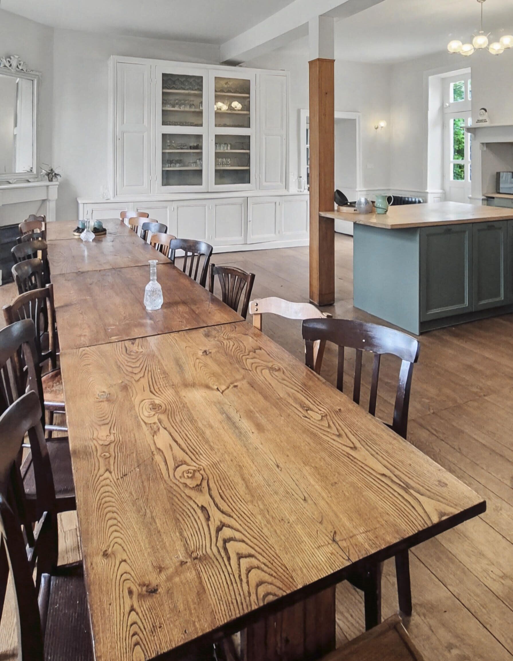 Un'ampia sala da pranzo con un lungo tavolo in legno e sedie, mobili da cucina bianchi con vetrate e un'isola centrale.