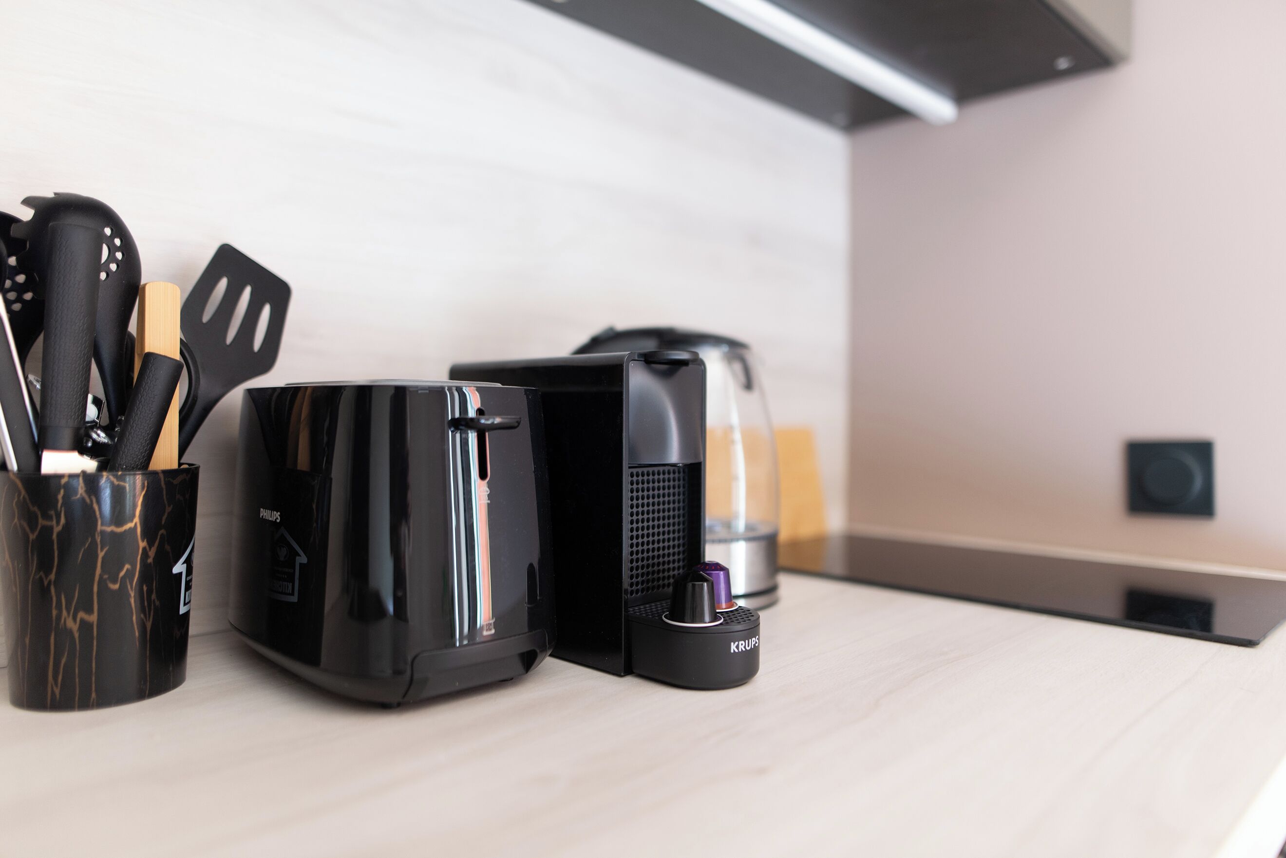 Une cuisine avec une machine à café noire, une bouilloire électrique noire et une cafetière noire. On y trouve une tasse noire contenant des ustensiles.