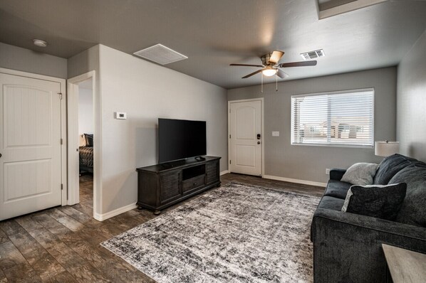 A living room with a flat-screen TV, dark wood entertainment center, gray sofa, patterned rug, and a ceiling fan.